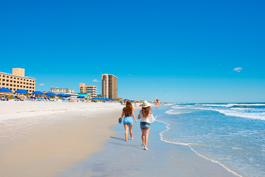 Two women stroll along the beach, enjoying the sun and waves, with footprints in the sand behind them.